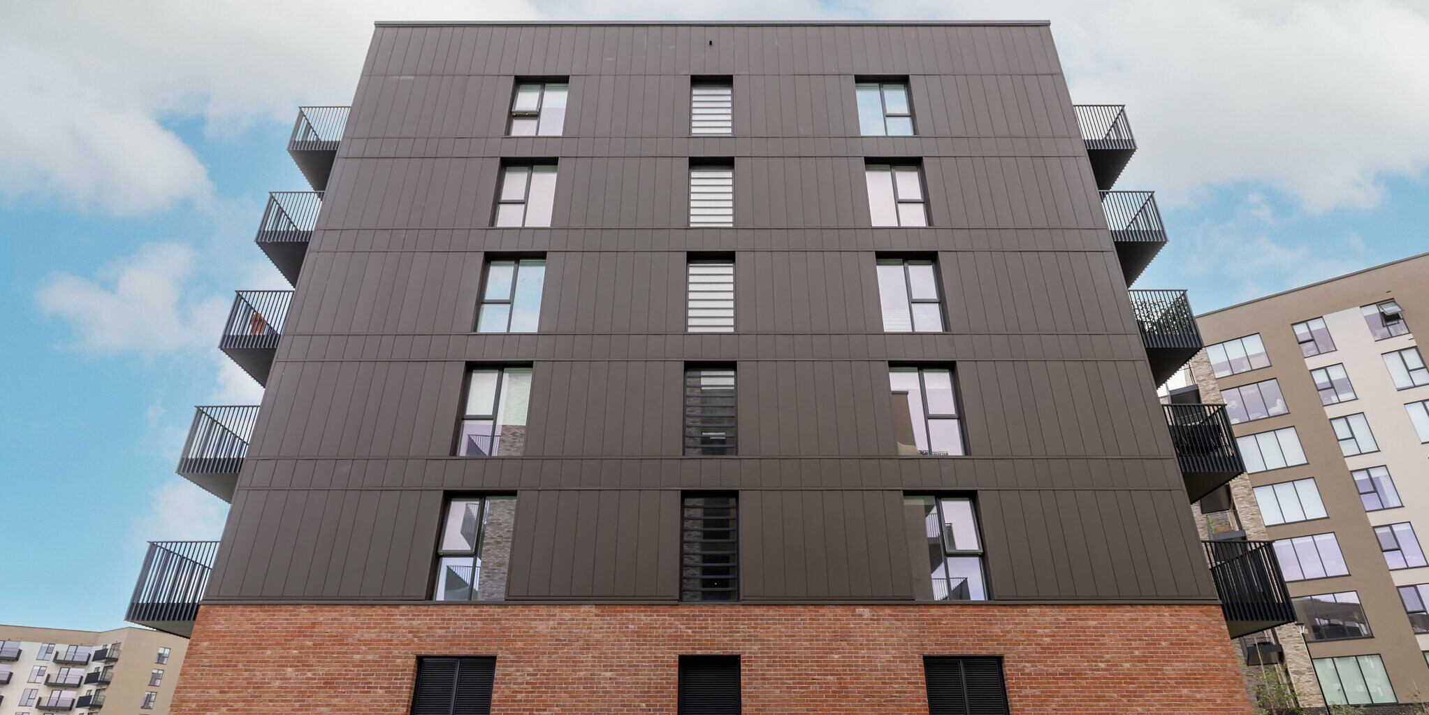 Froschperspektive der Citywest Apartments in Dublin. Die 700 m² vertikal verlegten PREFA Sidings in P.10 Braun verbinden sich mit der Ziegelfassade zu einer harmonischen und langlebigen Einheit. Die modernen Aluminiumpaneele von PREFA sorgen für Nachhaltigkeit und Design.