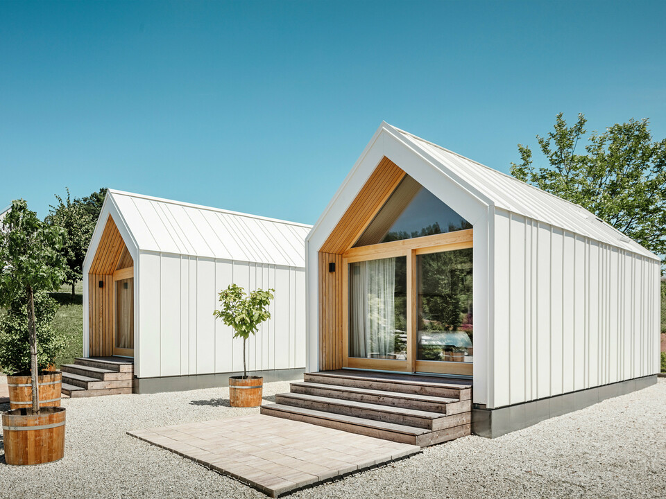 Zwei weiße Tiny Häuser der Ferienidylle 'Pri Momi' in Kančevci, Slowenien, gestaltet mit PREFALZ Dach- und Fassadenverkleidung in P.10 Reinweiß und naturbelassenem Holz. Die minimalistische Architektur der Gebäude wird durch den eleganten Materialmix aus Aluminium und Holz unterstrichen. Die großen Glasfronten an den Eingängen sowie die Holzstufen und umliegenden Bäume schaffen eine einladende und harmonische Atmosphäre. Die exklusive Unterkunft liegt inmitten einer malerischen Landschaft und wird speziell für Feiern genutzt.