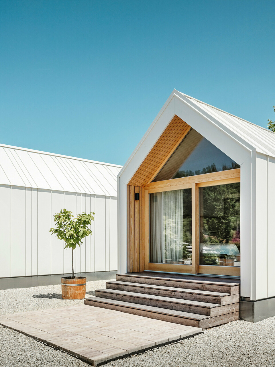 Zwei weiße Tiny Häuser der Ferienidylle 'Pri Momi' in Kančevci, Slowenien, gestaltet mit PREFALZ Dach- und Fassadenverkleidung in P.10 Reinweiß und naturbelassenem Holz. Die minimalistische Architektur der Gebäude wird durch den eleganten Materialmix aus Aluminium und Holz unterstrichen. Die großen Glasfronten an den Eingängen sowie die Holzstufen und umliegenden Bäume schaffen eine einladende und harmonische Atmosphäre. Die exklusive Unterkunft liegt inmitten einer malerischen Landschaft und wird speziell für Feiern genutzt.