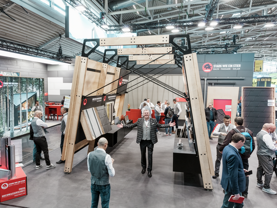 Lebendiges Treiben am PREFA Messestand auf der BAU Messe, mit Fokus auf eine große Produktpräsentationsstruktur, die Dach- und Fassadenelemente sowie Montageoptionen zeigt. Der Slogan „Stark wie ein Stier“ ist prominent sichtbar. Besucher und PREFA Mitarbeiter führen Fachgespräche. Der PREFA Geschäftsführer aus Deutschland, Karsten Köhler, macht eine einladende Geste und begrüßt alle Besucher auf dem Messestand.