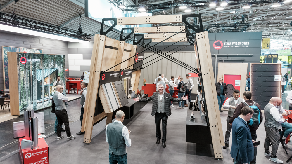 Lebendiges Treiben am PREFA Messestand auf der BAU Messe, mit Fokus auf eine große Produktpräsentationsstruktur, die Dach- und Fassadenelemente sowie Montageoptionen zeigt. Der Slogan „Stark wie ein Stier“ ist prominent sichtbar. Besucher und PREFA Mitarbeiter führen Fachgespräche. Der PREFA Geschäftsführer aus Deutschland, Karsten Köhler, macht eine einladende Geste und begrüßt alle Besucher auf dem Messestand.