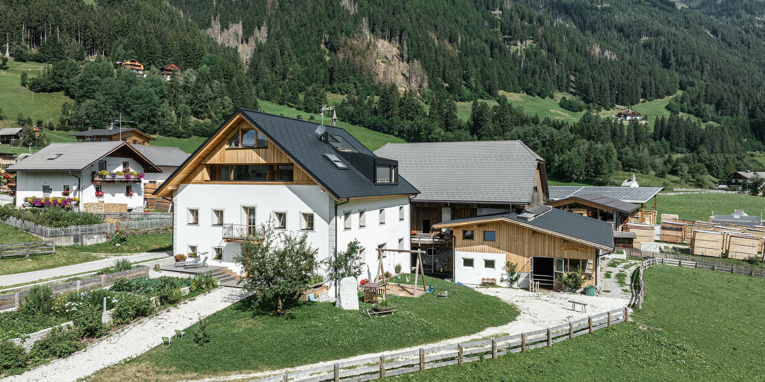 Der Außerwegerhof in Südtirol aus der Vogelperspektive mit einem PREFA-Langzeitdach in P.10 anthrazit. Die Ferienwohnungen liegen eingebettet in die grüne Landschaft des Antholzertals mit malerischen Wäldern und Bergen im Hintergrund. Die robuste PREFA DS.19 Dachschindel schützt die traditionellen Gebäude sowohl in den warmen Sommermonaten als auch bei eisigen Winterbedingungen.