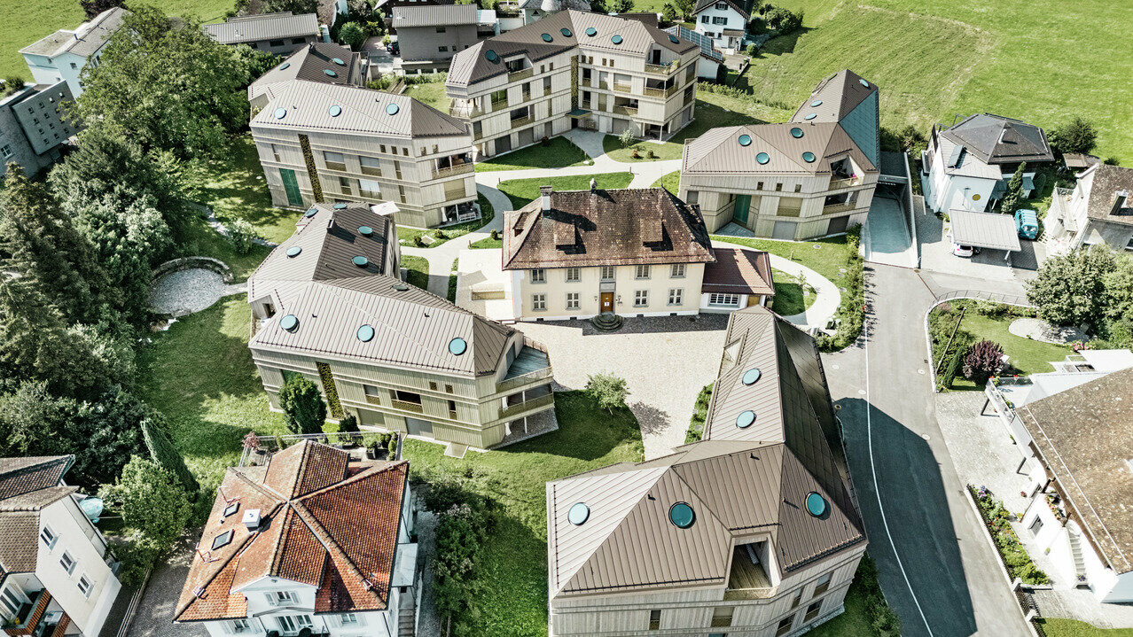 Luftaufnahme der modernen Wohnanlage in Liebwylen, Schweiz. Die Gebäude sind mit PREFALZ Aluminiumdächern in Braun gestaltet und verfügen über runde Dachfenster. Im Zentrum steht die historische Villa, umgeben von harmonisch angeordneten Neubauten, eingebettet in eine grüne Landschaft.