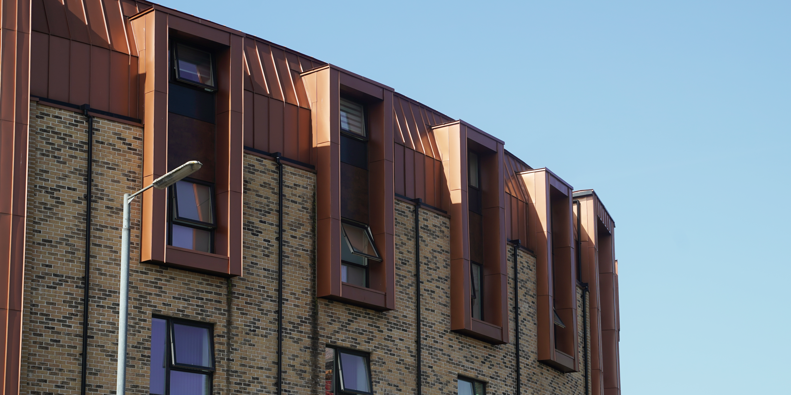 Die Fassade einer Wohnhausanlage in der Nethan Street in Glasgow, akzentuiert durch die geschmackvolle Verwendung von FALZONAL in Neukupfer. Das PREFA Aluminiumprodukt verleiht der Gebäudehülle durch seine Reflexion des Sonnenlichts ein lebendiges und einladendes Aussehen. Die Kombination der traditionellen Backsteinstruktur mit den modernen, kupferfarbenen Akzenten des FALZONAL vermittelt eine elegante Symbiose aus alt und neu und hebt die Baukunst in diesem städtischen Kontext hervor.