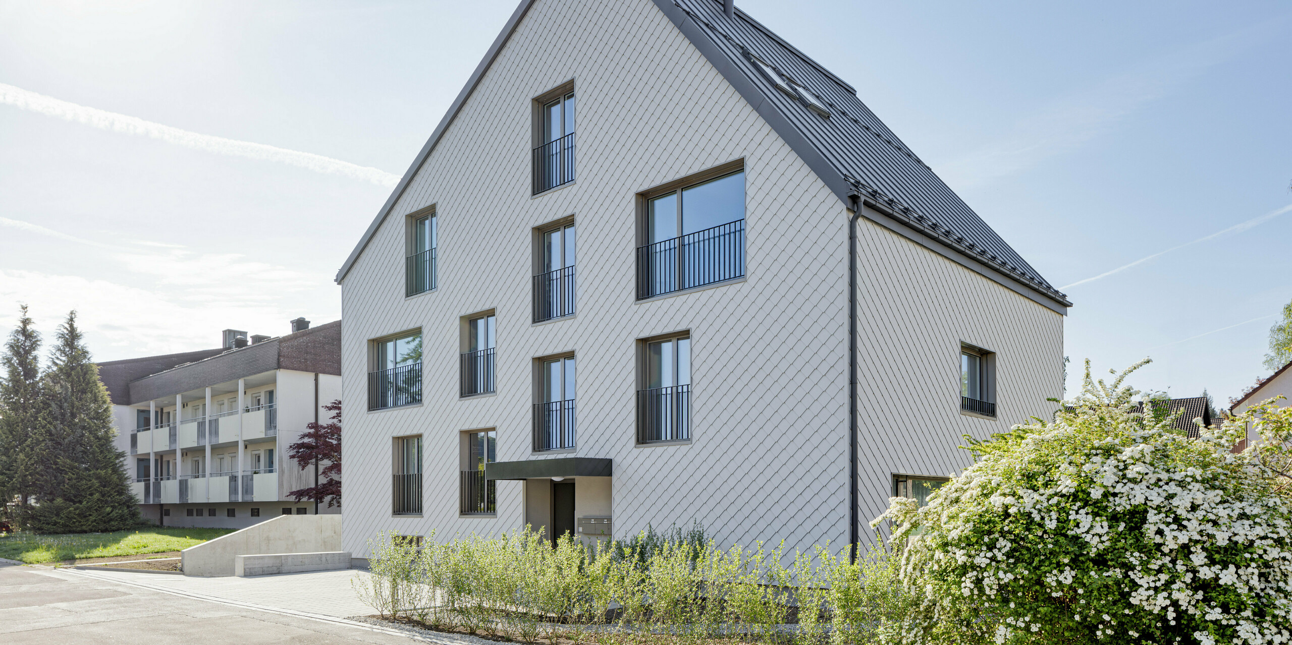 Modernes Mehrfamilienhaus in der Schweiz mit einem langlebigen PREFA Dach aus PREFALZ in P.10 Dunkelgrau und einer Fassade, verkleidet mit PREFA Wandrauten 20x20 in P.10 Prefaweiß. Das Objekt bietet eine zeitgemäße Architektur, die durch die Kombination von traditionellem Satteldach und minimalistischen Fassadenelementen besticht. Die klare Linienführung und das strukturierte Erscheinungsbild der Wandrauten sind harmonisch in eine gepflegte Umgebung mit üppigem Blütenstrauch integriert und zeigen die Vielseitigkeit von Aluminium als Baumaterial.