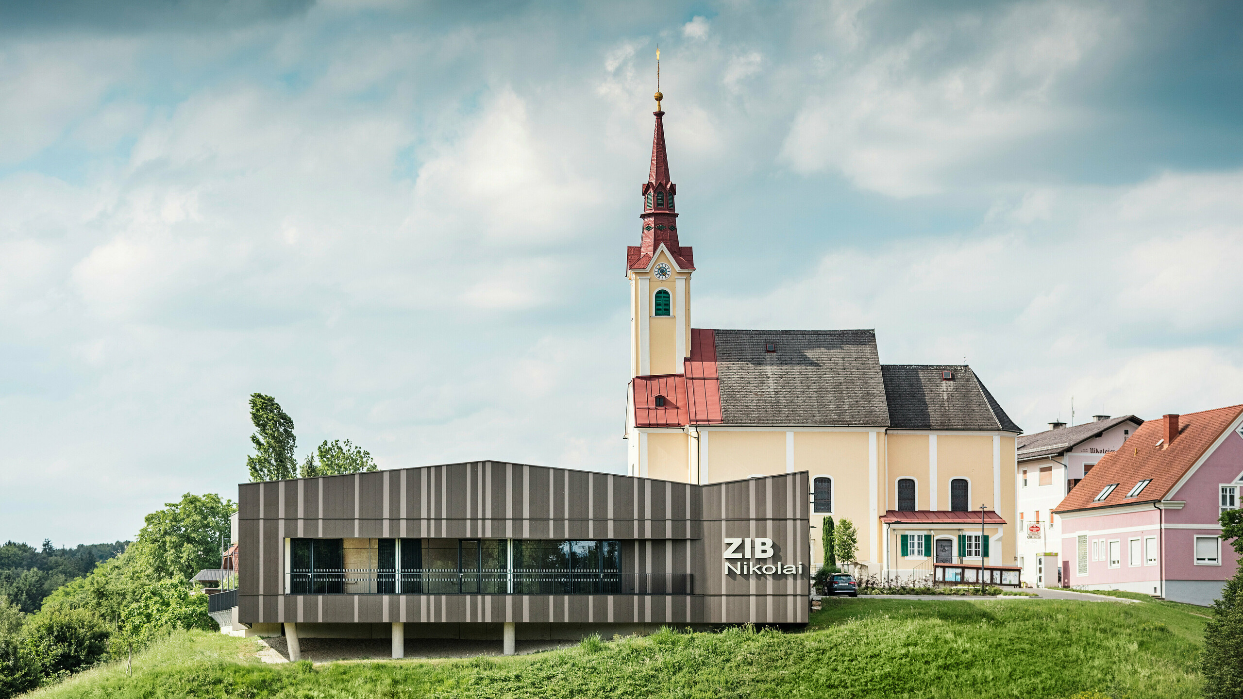 Das Veranstaltungszentrum ZIB Nikolai in St. Veit in der Südsteiermark mit einer modernen, dynamischen Aluminiumfassade in braun-beigegrauen Tönen, im Vordergrund eine grüne Wiese, im Hintergrund die Kirche von St. Nikolai mit gelber Fassade und rotem Dach.