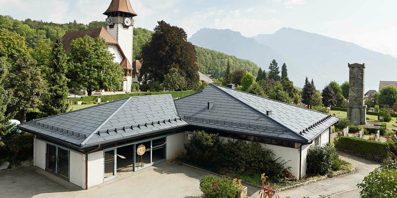 Aufbahrungshalle mit FX.12 Dachpaneelen von PREFA in Anthrazit, im Hintergrund steht eine Kirche und daneben befindet sich der Friedhof.