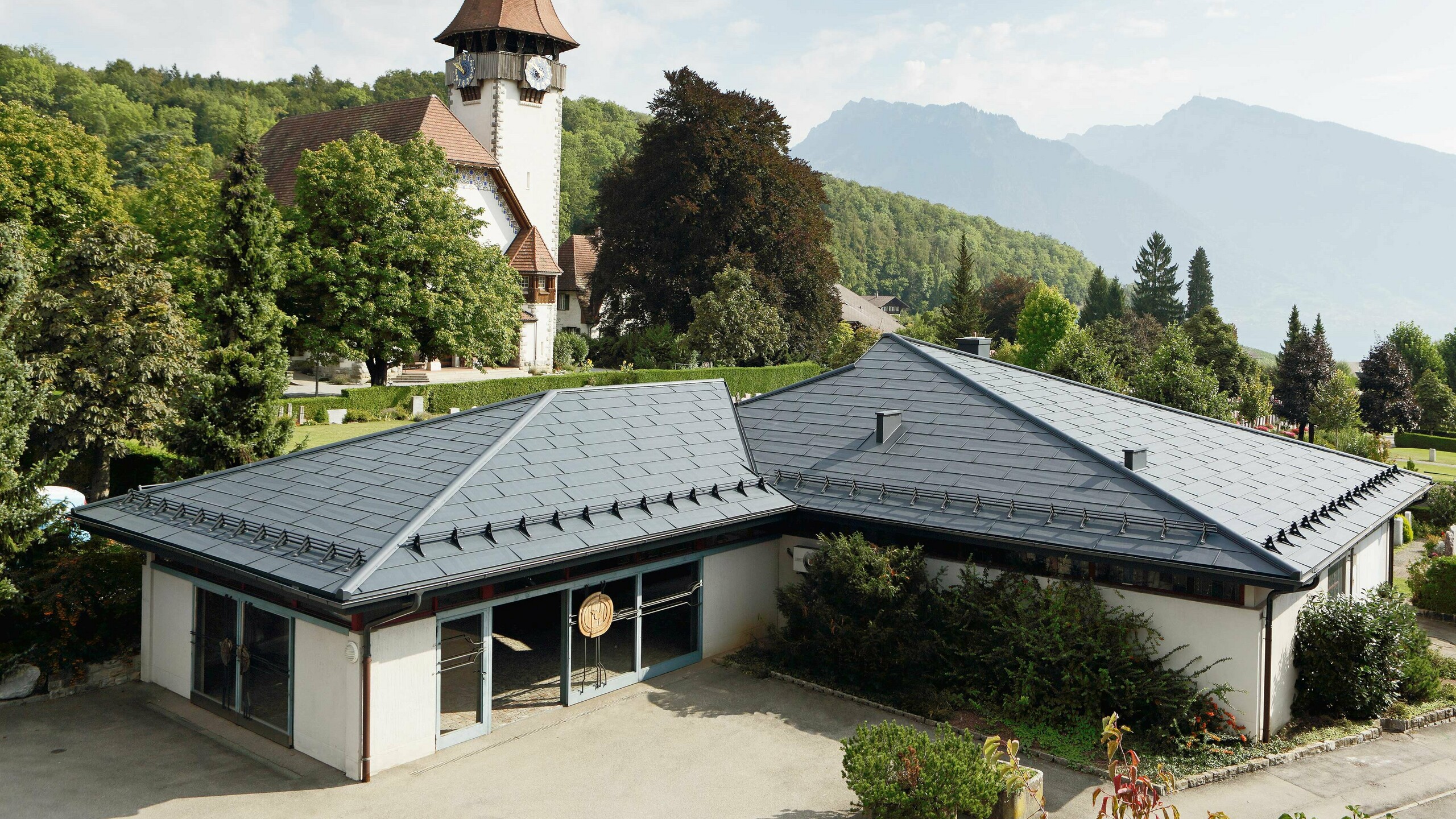 Aufbahrungshalle mit FX.12 Dachpaneelen von PREFA in Anthrazit, im Hintergrund steht eine Kirche und daneben befindet sich der Friedhof.