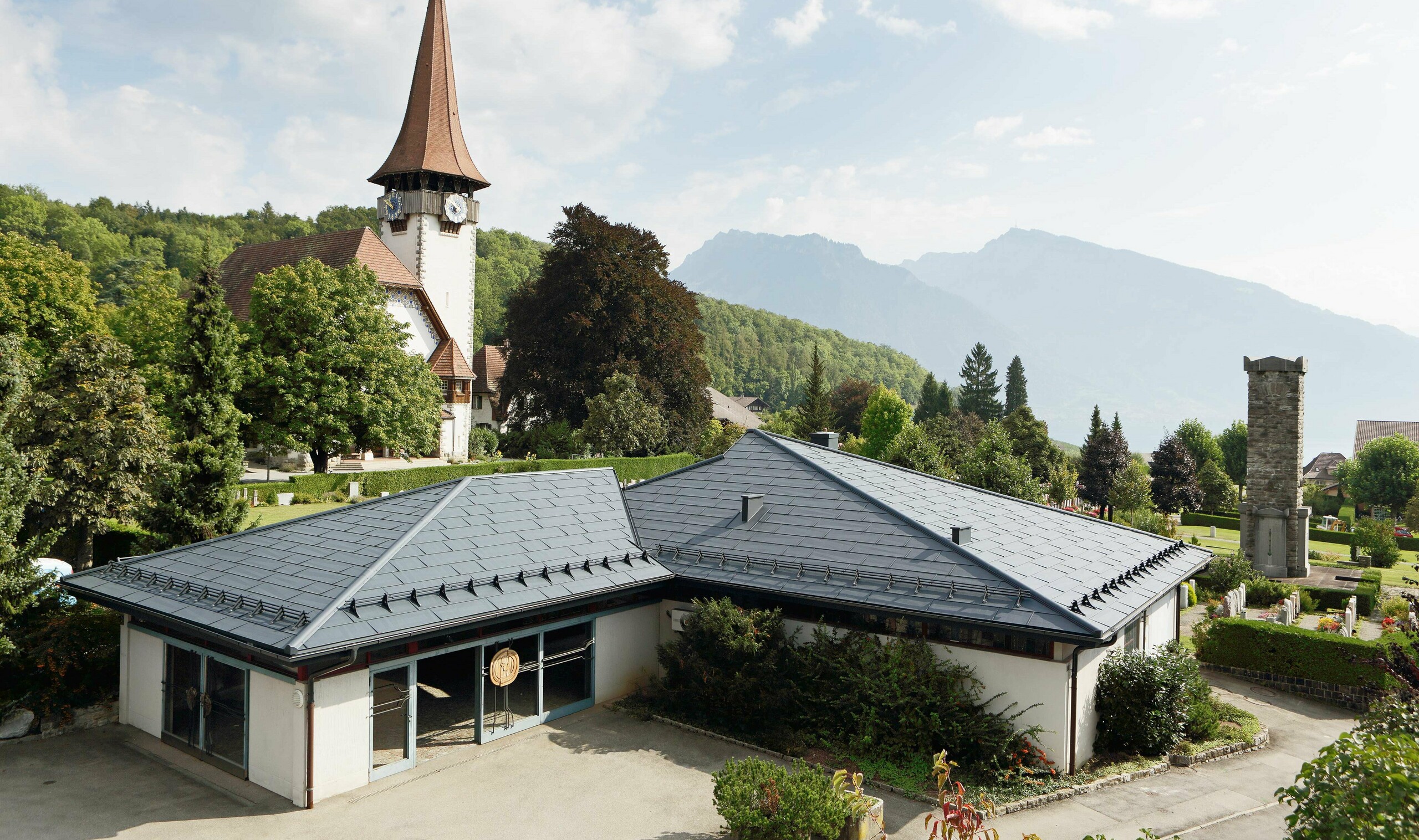 Aufbahrungshalle mit FX.12 Dachpaneelen von PREFA in Anthrazit, im Hintergrund steht eine Kirche und daneben befindet sich der Friedhof.