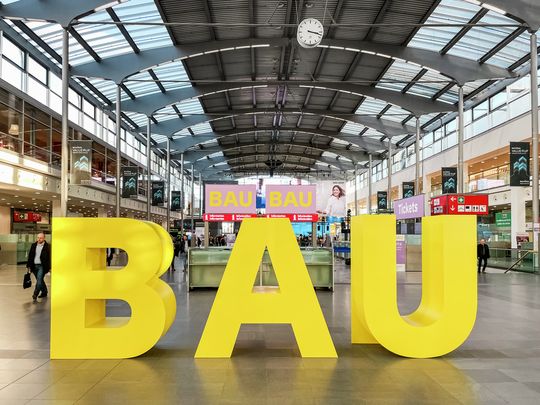 Innenaufnahme des Eingangsbereichs zur BAU Messe, der Weltleitmesse für Architektur, Materialien und Systeme. Im Vordergrund stehen große gelbe 3D-Buchstaben, die "BAU" buchstabieren. Der moderne, lichtdurchflutete Hallenbereich mit Glasdach und zahlreichen Hinweisschildern bildet den zentralen Ankunftspunkt für Besucher. Die Messe zieht Fachleute aus der Bau- und Architekturbranche aus aller Welt an.