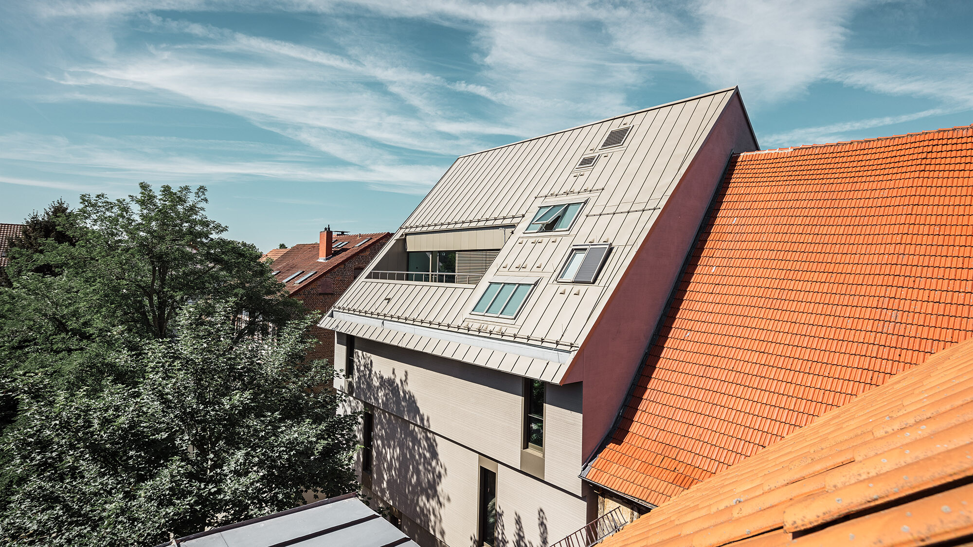 Ein seitlicher Blick auf das außergewöhnliche Dach: Links und rechts davon stehen Gebäude mit rostroten Biberschwanzziegeln, davor stehen Bäume.