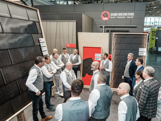 PREFA Team beim Gespräch auf der BAU Messe am Messestand, umgeben von innovativen Produkten für Dach, Fassade und Solar. Im Hintergrund sind Präsentationswände mit PREFA Dach- und Fassadenlösungen, darunter Solarmodule und Aluminium-Dachplatten, zu sehen. Das Motto „Stark wie ein Stier“ ist prominent dargestellt. Die Teammitglieder tragen einheitliche Firmenkleidung.