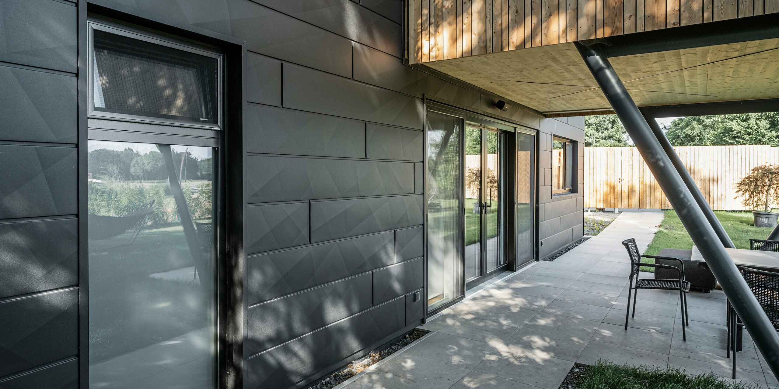 Blick auf die Außenterrasse des 'Woodstock Chalet 5', geschützt durch ein Vordach, das von schräg gestellten Stahlträgern gestützt wird. Die Terrasse wird von den dunklen, robusten PREFA Siding.X Fassadenelementen in Anthrazit eingerahmt, die eine elegante Optik und dauerhafte Qualität garantieren. Die Fassadenelemente fügen sich nahtlos in das moderne Design des Chalets ein und kontrastieren wirkungsvoll mit der natürlichen Holzfassade im oberen Bereich des Gebäudes.