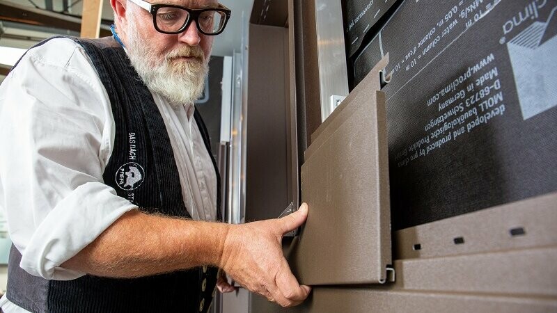 Zu sehen ist ein PREFA Techniker, der die entsprechend zugeschnittene PREFA Siding-Paneele in Braun unter dem Fenster montiert.