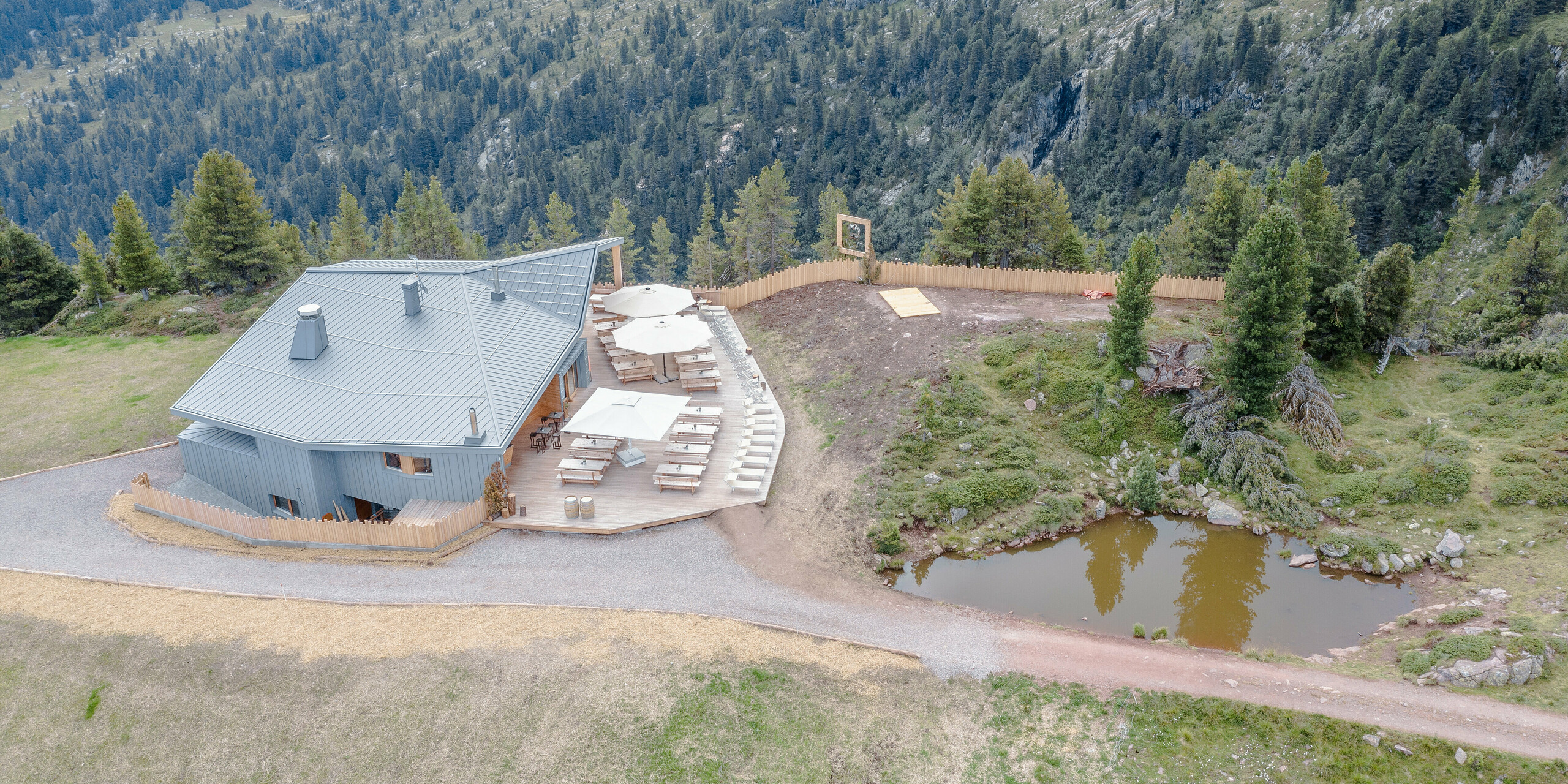 Luftbild des Ristorante Busabelle inmitten einer bergigen Landschaft. Die Architektur des Restaurants zeichnet sich durch eine moderne, hellgraue Gebäudehülle aus Blech aus. Das PREFALZ Dach- und Fassadensystem fügt sich geschmeidig in die natürliche Umgebung ein. Neben dem Gebäude befindet sich eine attraktiv gestaltete Terrasse mit Sonnenschirmen und Holztischen, die zum Entspannen einladen. Ein kleiner natürlicher Teich neben dem Restaurant fügt zusätzliche Idylle und Harmonie zum Standort hinzu.