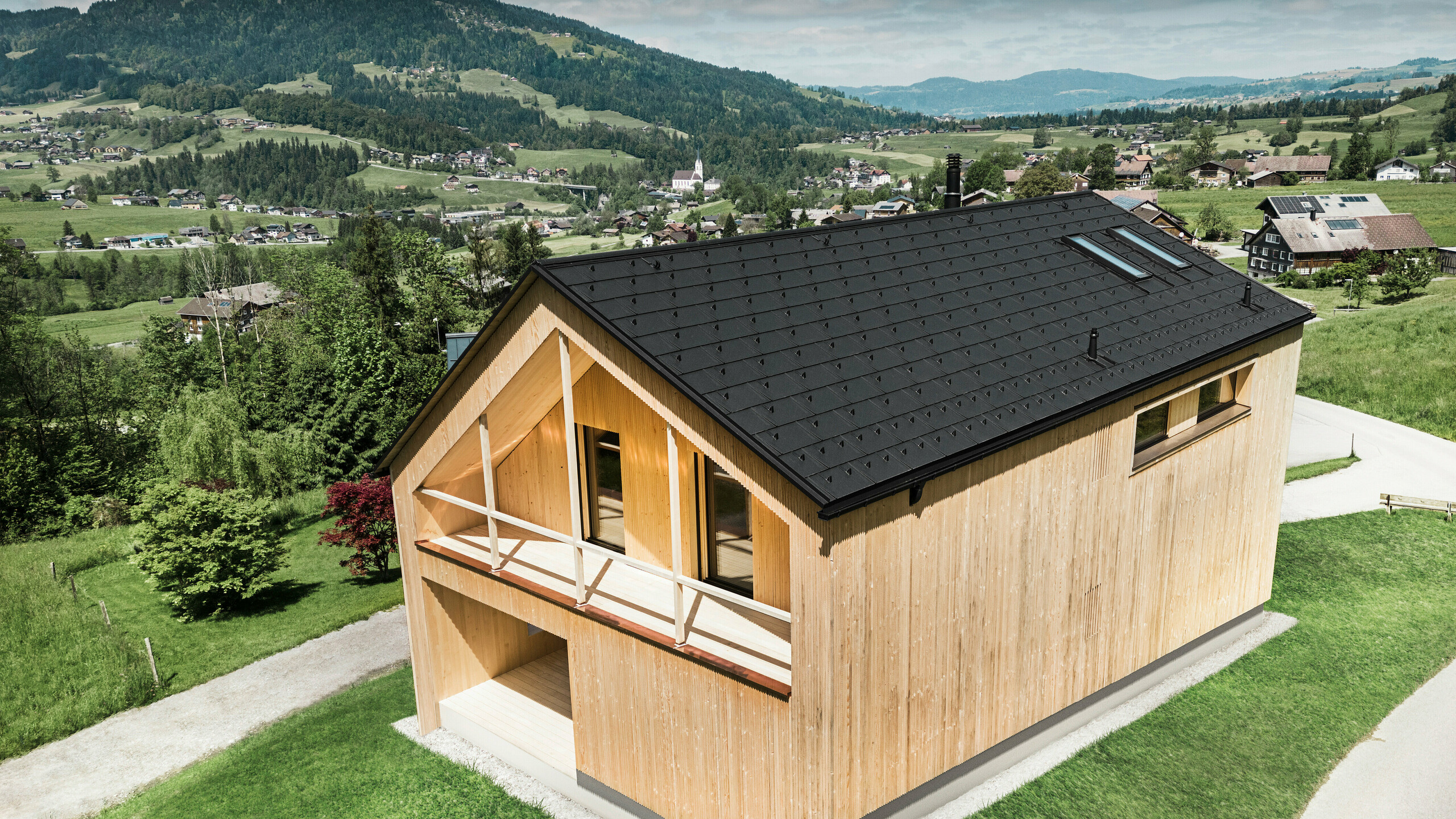 Dieses Foto zeigt ein ökologisches Vorzeigehaus in Egg, das sich mit seiner PREFA Solardachplatte und den schwarzen Dachplatten R.16 gegen die malerische Kulisse eines österreichischen Dorfes abhebt. Die Solarelemente sind nahtlos in die Dachstruktur eingebettet und liefern Energie aus einer erneuerbaren Energiequelle, während das natürliche Holz der Fassade und die sattgrüne Umgebung eine Verbindung von Nachhaltigkeit und Lokalität darstellen. Der Blick auf die umliegende dörfliche Struktur mit traditionellen Bauten im Hintergrund verleiht dem Bild eine zusätzliche Dimension und betont die gelungene Integration moderner, umweltfreundlicher Bauweisen in gewachsene Landschaften.