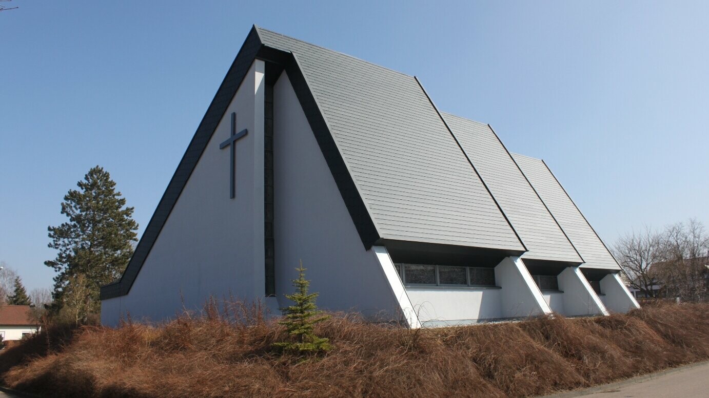 Die Kirche in Bettringen ist mit PREFA Dachschindeln in P.10 Anthrazit eingedeckt. Das moderne Gebäude zeichnet sich durch seine markanten, schrägen Dachflächen und die klare, geometrische Architektur aus. Auf der Vorderseite des Gebäudes ist ein großes Kreuz zu sehen.