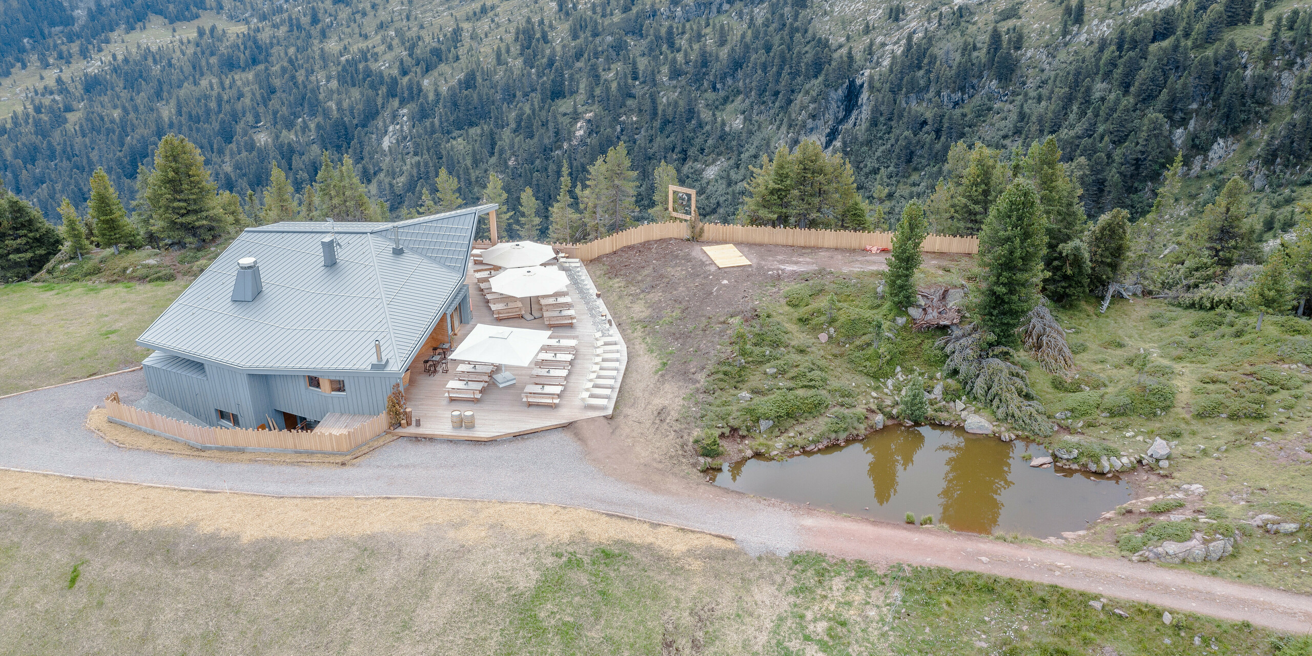 Luftbild des Ristorante Busabelle inmitten einer bergigen Landschaft. Die Architektur des Restaurants zeichnet sich durch eine moderne, hellgraue Gebäudehülle aus Blech aus. Das PREFALZ Dach- und Fassadensystem fügt sich geschmeidig in die natürliche Umgebung ein. Neben dem Gebäude befindet sich eine attraktiv gestaltete Terrasse mit Sonnenschirmen und Holztischen, die zum Entspannen einladen. Ein kleiner natürlicher Teich neben dem Restaurant fügt zusätzliche Idylle und Harmonie zum Standort hinzu.