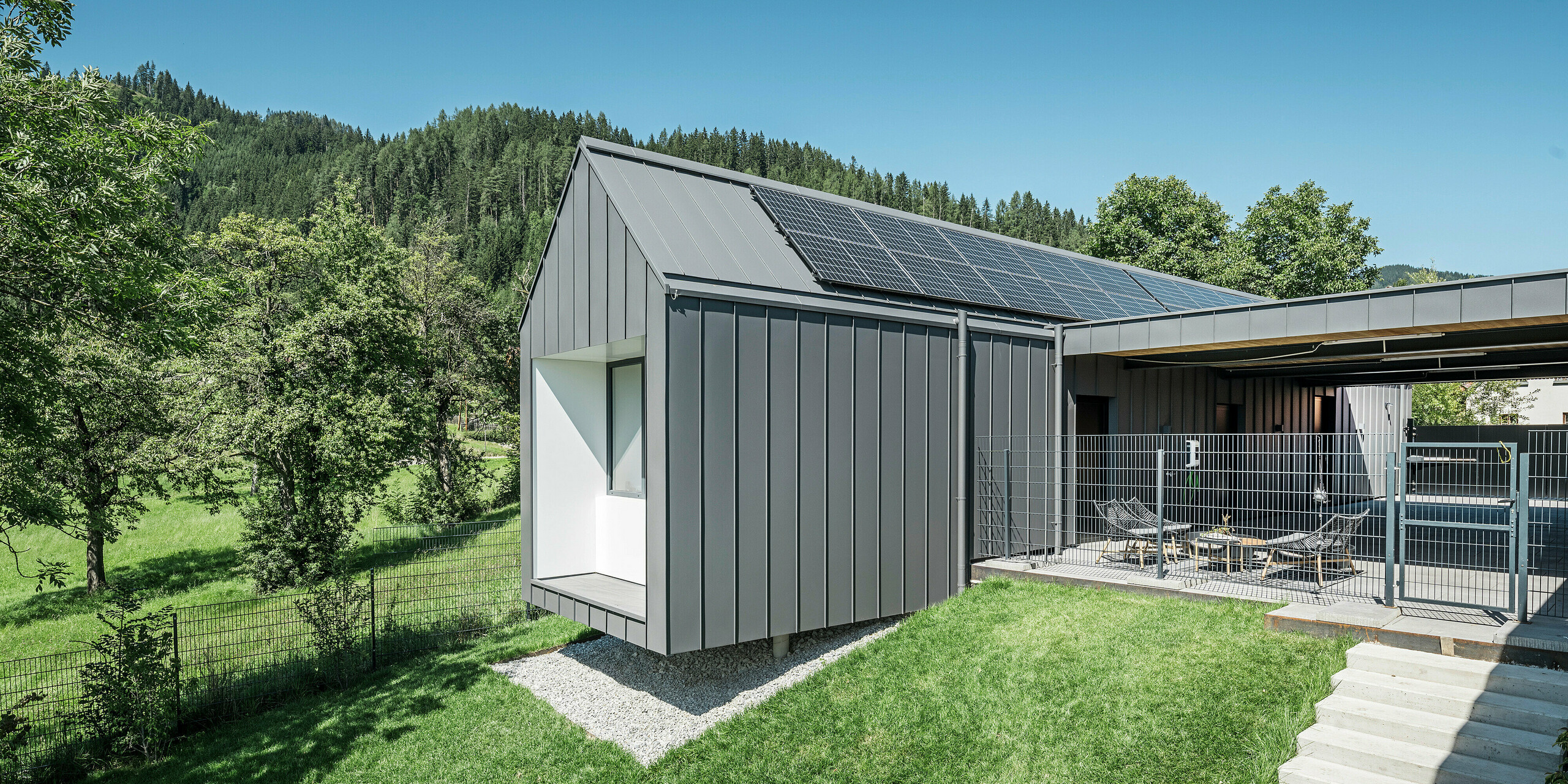 Harmonische Rückansicht eines modernen Einfamilienhauses in Pogier, Österreich, mit dem PREFALZ Dach- und Fassadensystem dunkelgrau P.10, integriert in die idyllische Waldlandschaft. Die Kombination aus dunkelgrauer Aluminiumverkleidung und natürlicher Umgebung unterstreicht das ökologische Konzept des Hauses, das durch eine effiziente Photovoltaikanlage auf dem Dach ergänzt wird. Der eingezäunte Außenbereich mit gemütlichen Sitzgelegenheiten lädt zum Verweilen ein, um die Ruhe und Frische des umliegenden Waldes zu genießen.