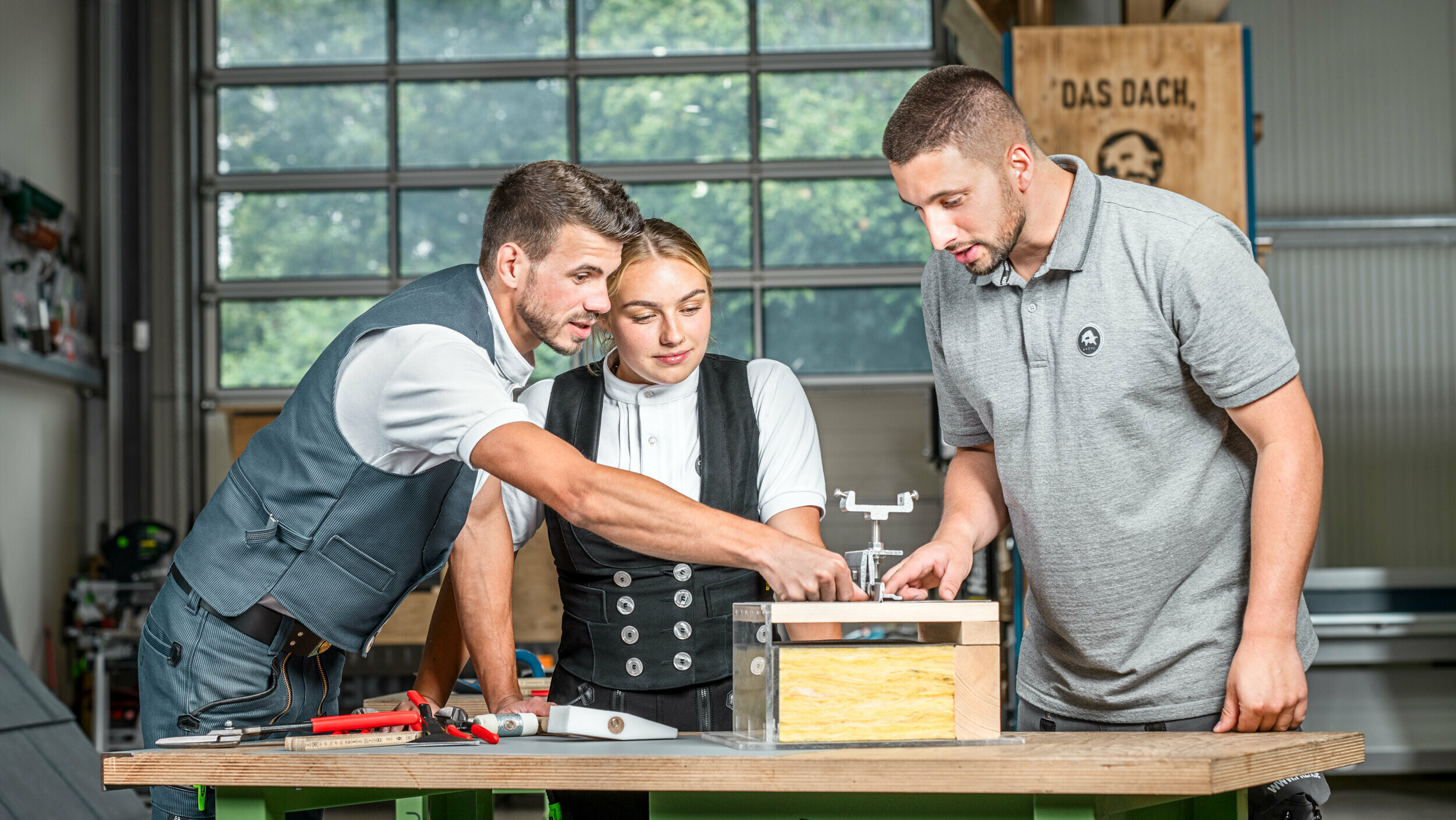 PREFA Anwendungstechniker Julian Eickhoff, Marvin Bürger von Bürger Falztechnik und Dachdeckermeisterin Fabienne Ellermeier schauen sich gemeinsam ein Lehrmodell in der PREFA Academy Rheinbach an. 