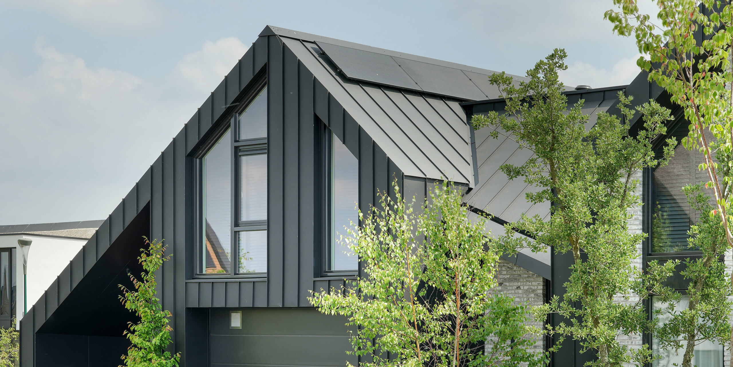 Detail eines modernen Einfamilienhauses in Veenendaal, Niederlande, das in einer natürlichen Umgebung gezeigt wird. Das Gebäude zeichnet sich durch sein schwarzes PREFALZ Spitzdach aus. Die schwarze Aluminium-Verkleidung des Gebäudes bildet einen auffälligen Kontrast zu den großen Fenstern und der hellgrauen Naturstein-Fassade. Die abwechslungsreiche Mischung aus dunklen und hellen Oberflächen schafft ein spannendes Nebeneinander, das durch die umgebende Grünfläche noch akzentuiert wird.