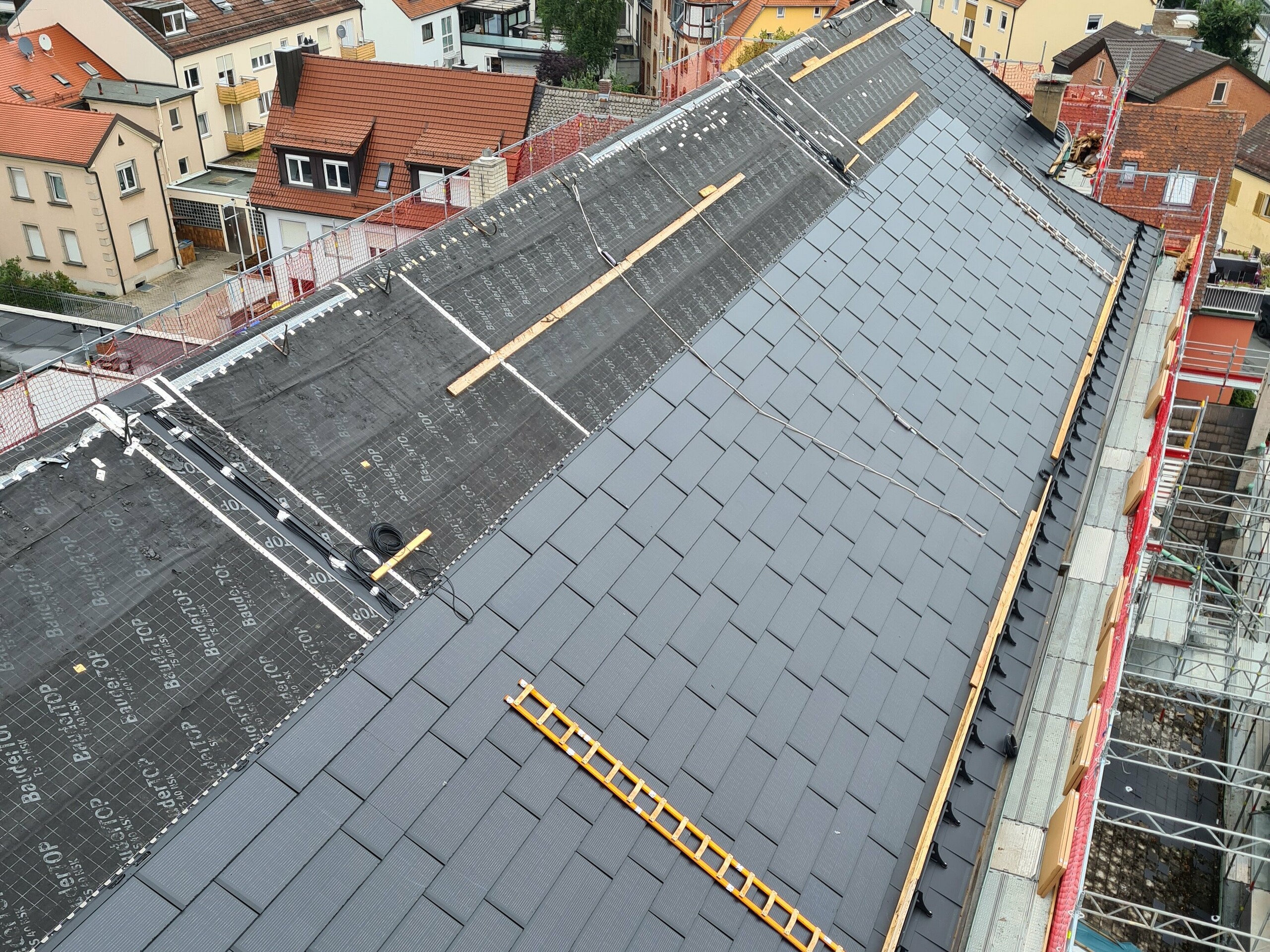 Vogelperspektive auf das teilweise neu sanierte Dach der St.-Karl-Borromäus-Kirche, das mit großen PREFA Solardachplatten ausgestattet ist. Die Platten sind gleichmäßig auf dem gesamten Dach verlegt und haben eine dunkle, glänzende Oberfläche. Die Architektur der Kirche ist im Hintergrund sichtbar, einschließlich der charakteristischen Dachform und des Turms. Die Solardachplatten sind präzise montiert und passen sich optisch gut in das denkmalgeschützte Gebäude ein.