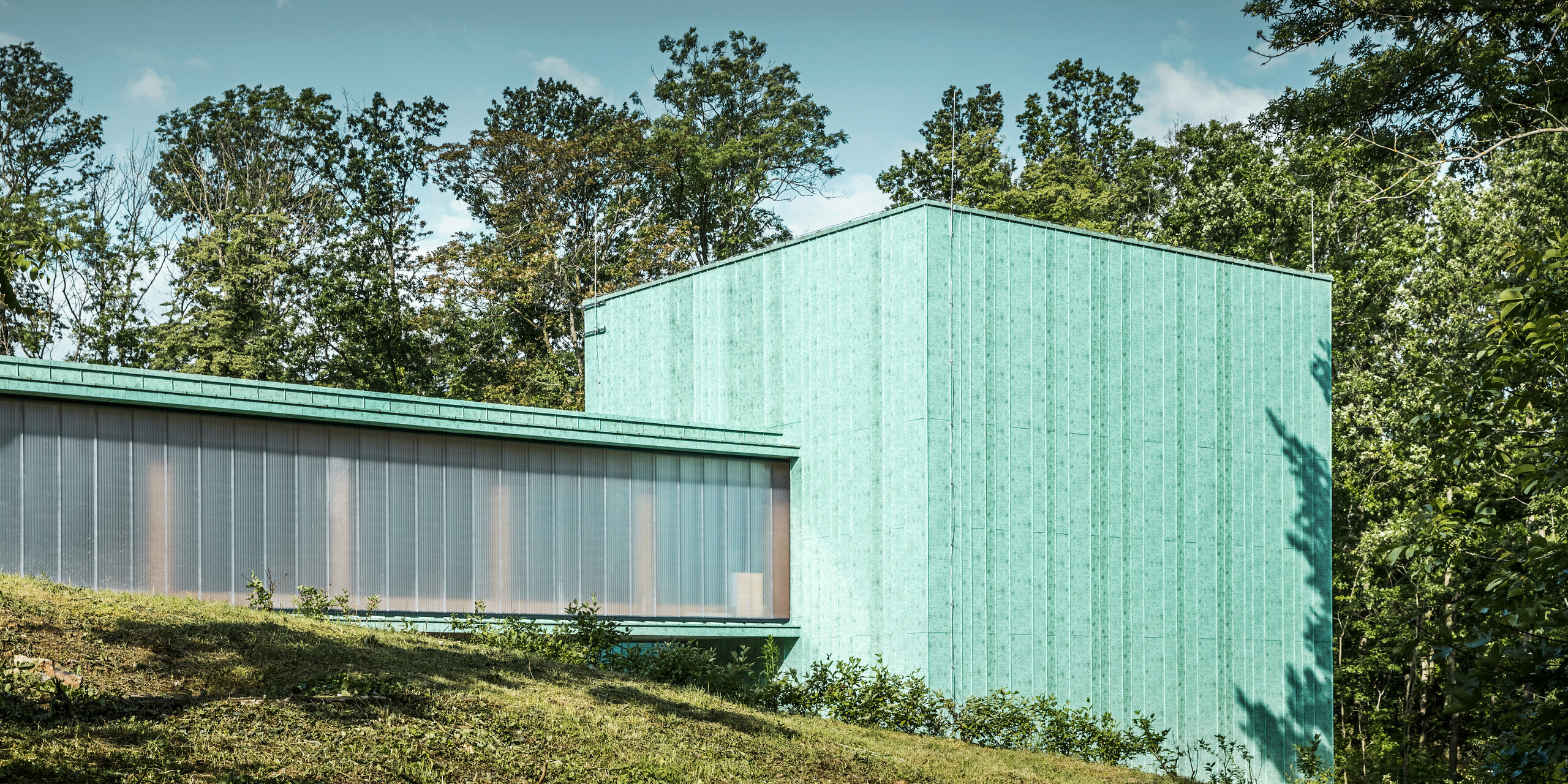 Das Foto präsentiert das Zero Magnetic Field Laboratory in Fertöboz, Ungarn. Das einzigartige Gebäude ist mit einer PREFA Aluminiumfassade in Patinagrün verkleidet. Das Fassadensystem PREFALZ wurde in einer vertikalen Struktur verlegt. Das patinagrüne Bauwerk fügt sich nahtlos in die umgebende Landschaft ein. Der spezielle Grünton der Fassade harmoniert mit dem Grün der Bäume und dem Himmel, was die Langlebigkeit von Aluminium als Baumaterial unterstreicht. Der Schattenwurf auf der Fassade deutet auf die Interaktion mit der natürlichen Umgebung hin und verleiht dem Bild eine zusätzliche Tiefe. Das Bild zeigt zudem auf der linken Seite einen Durchgang zu einem weiteren Gebäudeteil. Der Tunnel wurde in einer leicht transparenten Verglasung an den Seiten ausgestattet. 