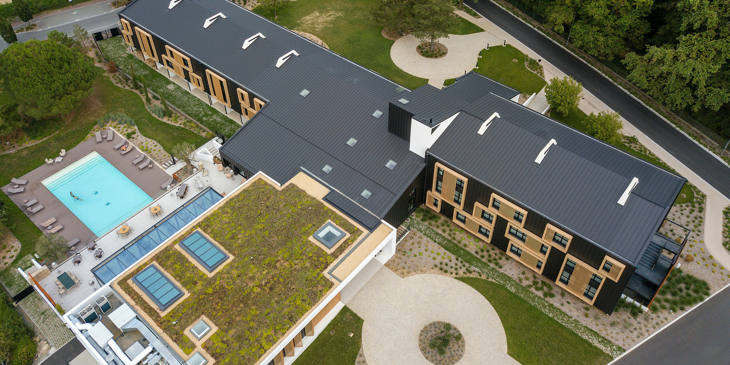 Luftaufnahme auf den Eingangsbereich des Hôtel Parc du Landreau in Les Herbiers, Frankreich. Das Gebäude wurde mit dem PREFALZ Dach- und Fassadensystem in Schwarzgrau verkleidet. Die markanten Holzrahmen um die Fenster ergänzen die moderne Aluminiumverkleidung. Der Außenbereich mit Swimmingpool und teilweise begrüntem Dach spiegelt die nachhaltige Bauweise des Hotels wider.