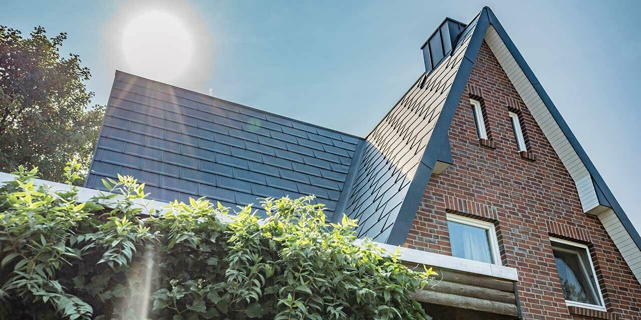 Satteldach mit der schlichten PREFA Alu-Dachplatte R.16 in Anthrazit eingedeckt. Die Fassade ist eine rustikale Klinkerfassade, hinter dem Haus geht gerade die Sonne auf.