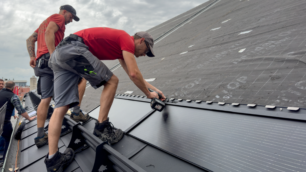 Ein Verarbeiter befestigt eine Reihe mit großen, schwarzen PREFA Solardachplatten auf dem Dach der St. Karl Borromäus Kirche in Nürnberg. Der Schrauber ist auf der Oberfläche der Solardachplatte positioniert. Im Hintergrund ist ein Teil des Daches sowie ein unscharfer Bereich zu erkennen, der auf eine größere Dachfläche hinweist.