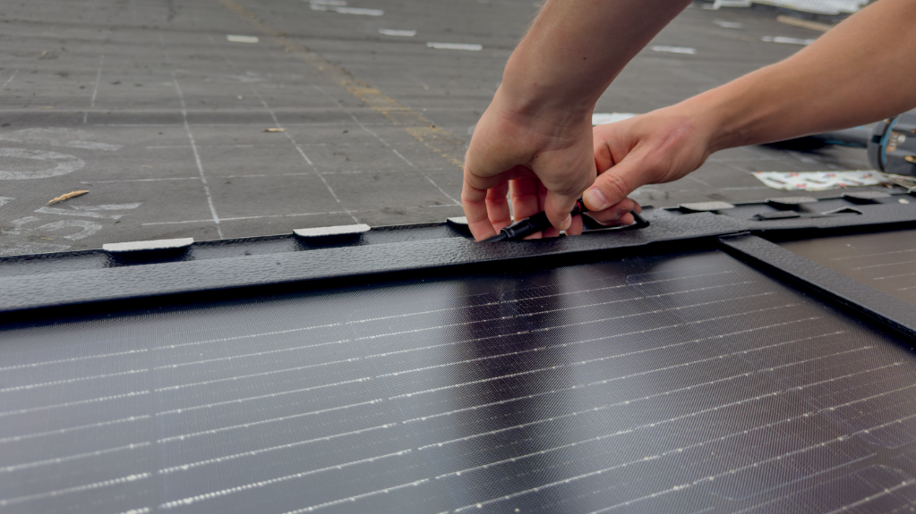 Nahaufnahme der Installation einer großen PREFA Solardachplatte auf dem Dach der St. Karl Borromäus Kirche in Nürnberg (Bayern). Die glänzenden, rechteckigen Platten sind sauber nebeneinander aufgereiht und haben eine schwarze Oberfläche. Die Hände des Handwerkers sind im rechten Teil des Bildes zu sehen. Er schließt die Kabel der Solardachplatte an. Der Hintergrund zeigt das bereits teilweise verlegte und vorbereitete Dach. Die Szene findet unter freiem Himmel statt, was auf die Außenmontage der Solarmodule hinweist.