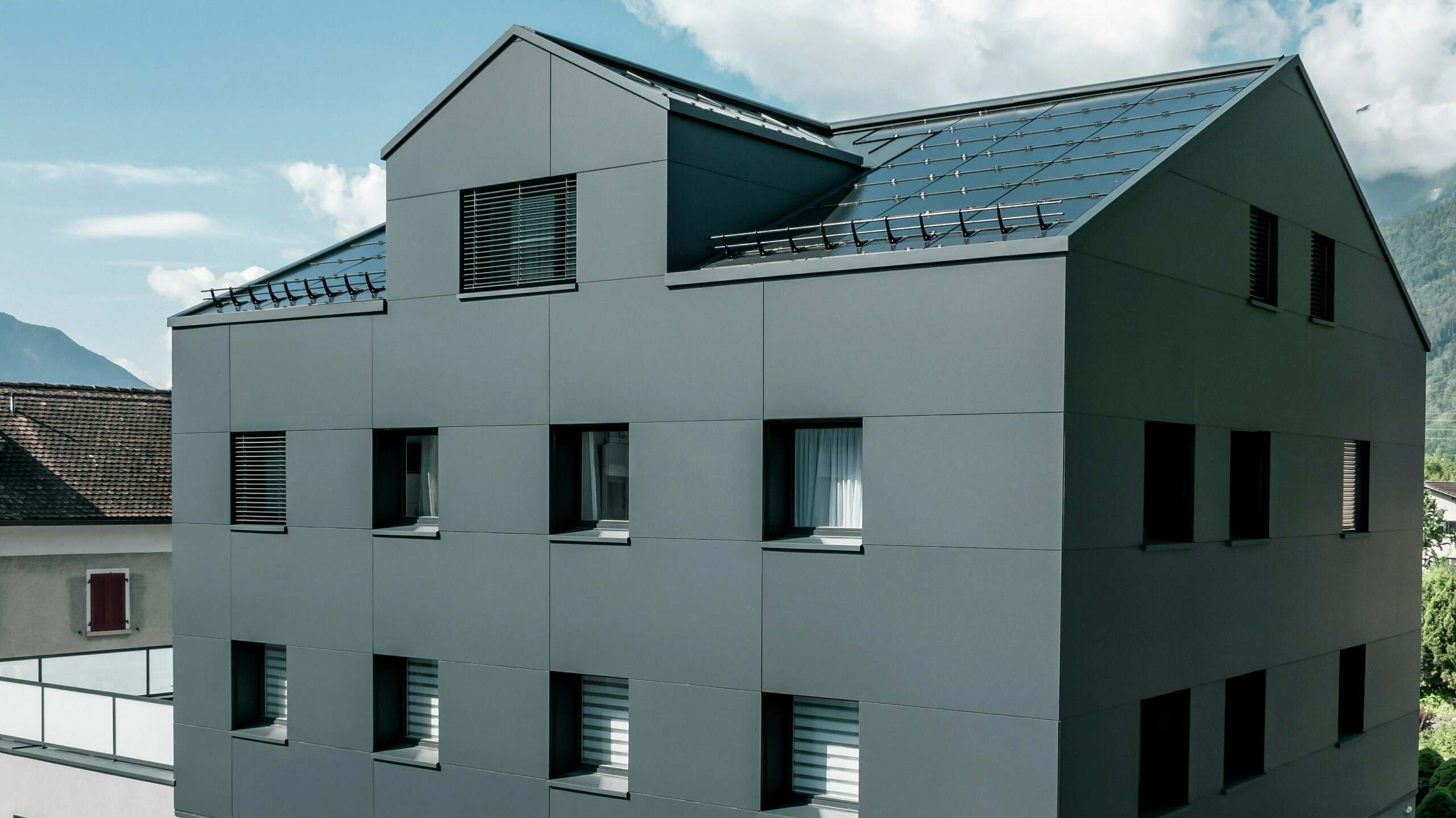 Modernisiertes Wohn- und Geschäftsgebäude nach der Fassadensanierung mit PREFABOND Aluminium Verbundplatten in Anthrazit. Die klare, elegante Linienführung der neuen Fassade verleiht dem Gebäude ein modernes Erscheinungsbild. Im Erdgeschoss befindet sich das Architekturbüro Voser. Die Umgebung mit Bäumen und Bergen rundet die harmonische Optik ab.