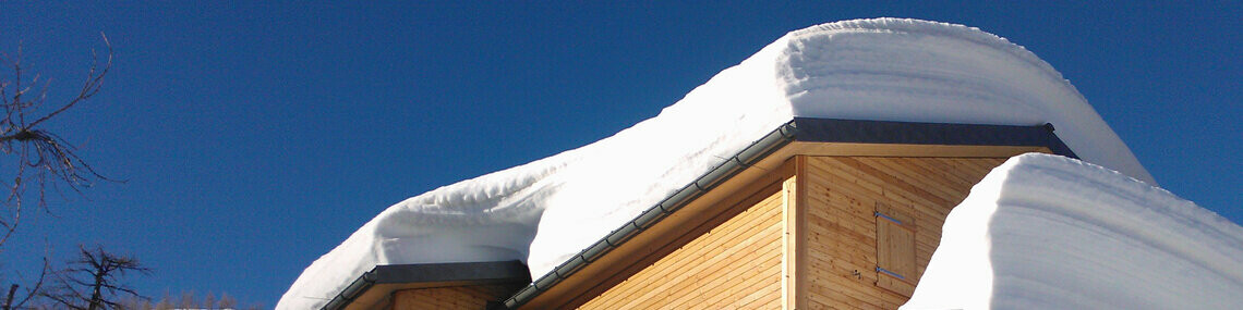 Das Bild zeigt die Hütte Capanna Buffaloraim im Winter, die von dicken Schneewechten am Dach bedeckt ist. Das Bild weist auf die Gefahren durch abrutschende Schneelawinen hin. Das Dach der Berghütte ist mit robusten PREFA Dachpaneelen FX.12-gedeckt, welche besonders für extreme Wetterbedingungen geeignet sind. Trotz der massiven Schneelast bleibt die Dachkonstruktion stabil und sicher. Dies zeigt die hohe Belastbarkeit und Qualität der PREFA Produkte. Die Berghütte selbst besteht aus Holz und fügt sich harmonisch in die winterliche Landschaft ein.