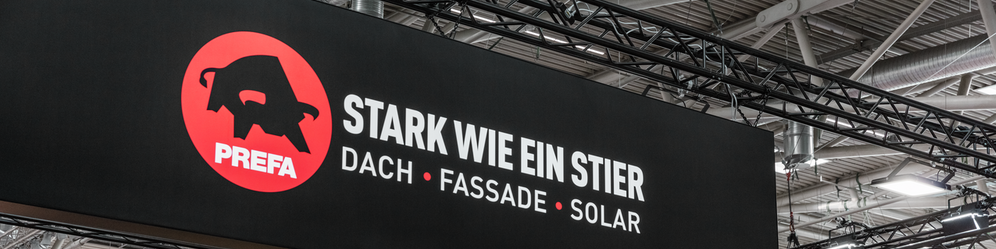 Großes PREFA Logo mit dem Slogan „Stark wie ein Stier – Dach, Fassade, Solar“ auf einer hängenden Werbetafel am Messestand auf der BAU Messe. Im Hintergrund sind Visualisierungen moderner Architekturprojekte mit PREFA Fassadenlösungen sowie Teile anderer Messestände zu sehen.