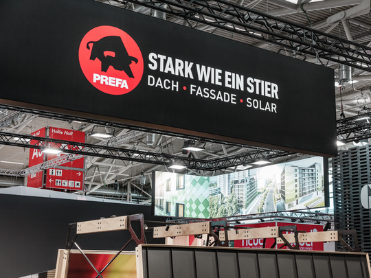 Großes PREFA Logo mit dem Slogan „Stark wie ein Stier – Dach, Fassade, Solar“ auf einer hängenden Werbetafel am Messestand auf der BAU Messe. Im Hintergrund sind Visualisierungen moderner Architekturprojekte mit PREFA Fassadenlösungen sowie Teile anderer Messestände zu sehen.