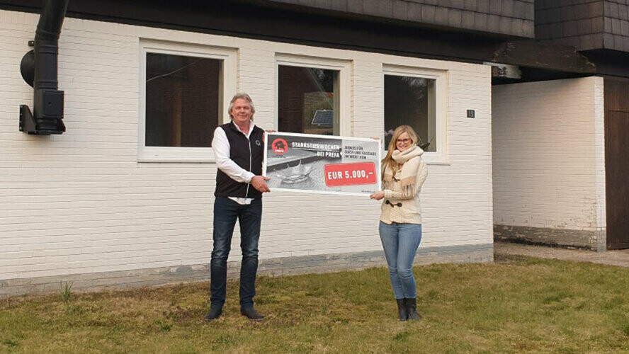 Ein Mann und eine Frau stehen vor ihrem Haus und lächeln und halten einen Scheck in der Hand.