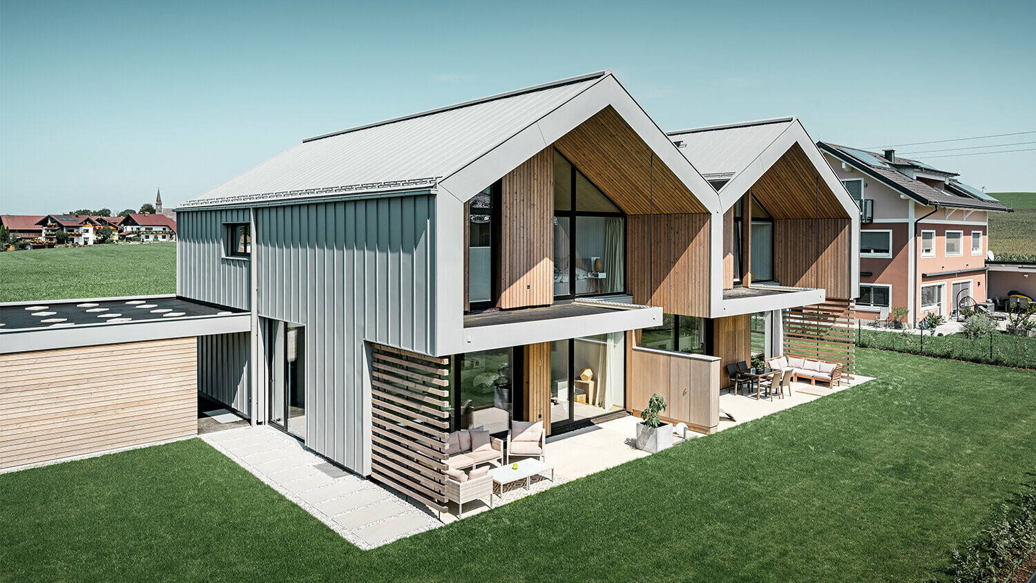 Ein modernes Doppelhaus mit einer Gebäudehülle aus Aluminium. Das Blechdach sowie die Blechfassade bestehen aus PREFALZ in Patinagrau. Die helle Stehfalzdeckung kombiniert mit den Holzelementen fügt sich harmonisch in die Umgebung ein. Die Dachentwässerung erfolgt über das PREFA Quadratrohr und ist ebenfalls in der Farbe Patinagrau gehalten. 