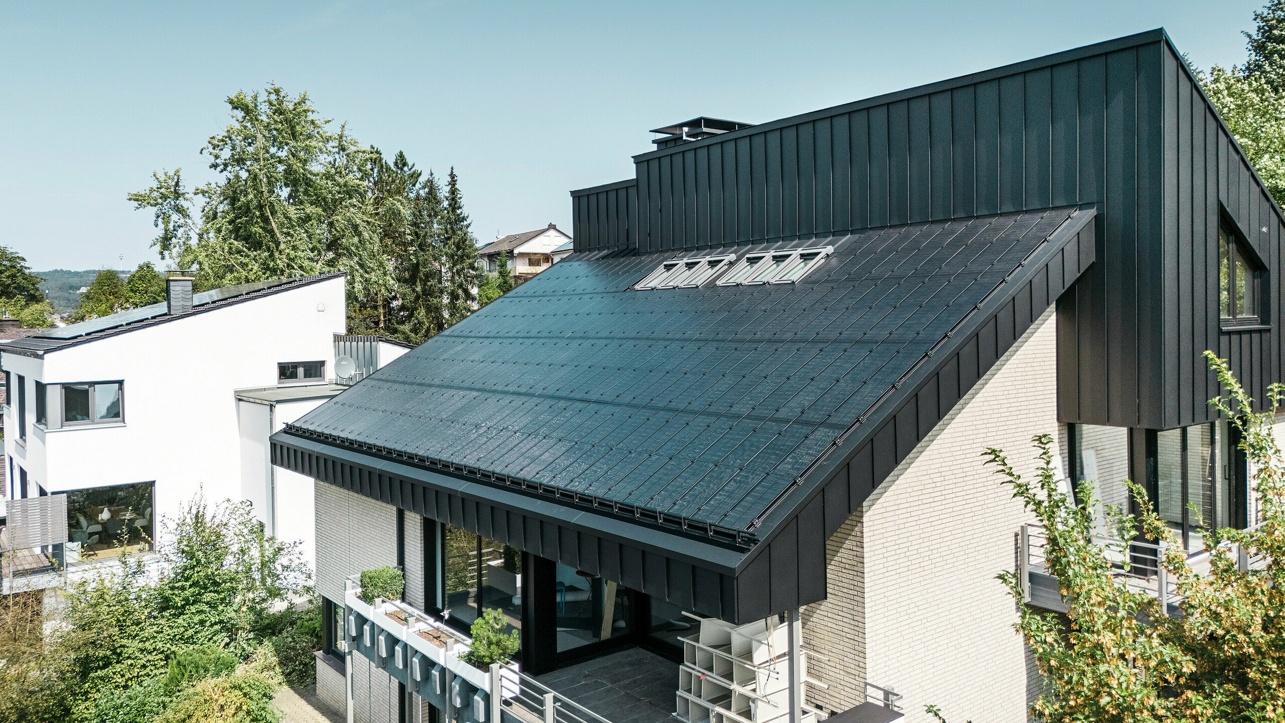 Moderne Architektur eines Einfamilienhauses in Meschede, Deutschland, mit einem PREFALZ Aluminiumdach in P.10 Schwarz und integrierten Glas/Glas PV-Modulen. Das versetzte Pultdach verbindet zeitloses Design mit nachhaltiger Photovoltaik-Technologie. Die innovativen PREFALZ Solarmodule liefern eine Leistung von 18 kWp. Die rahmenlosen Module und die elegante Aluminiumverkleidung von Fassade und Dach bilden eine ästhetische und funktionale Einheit, die sich perfekt in das grüne Wohnumfeld einfügt.